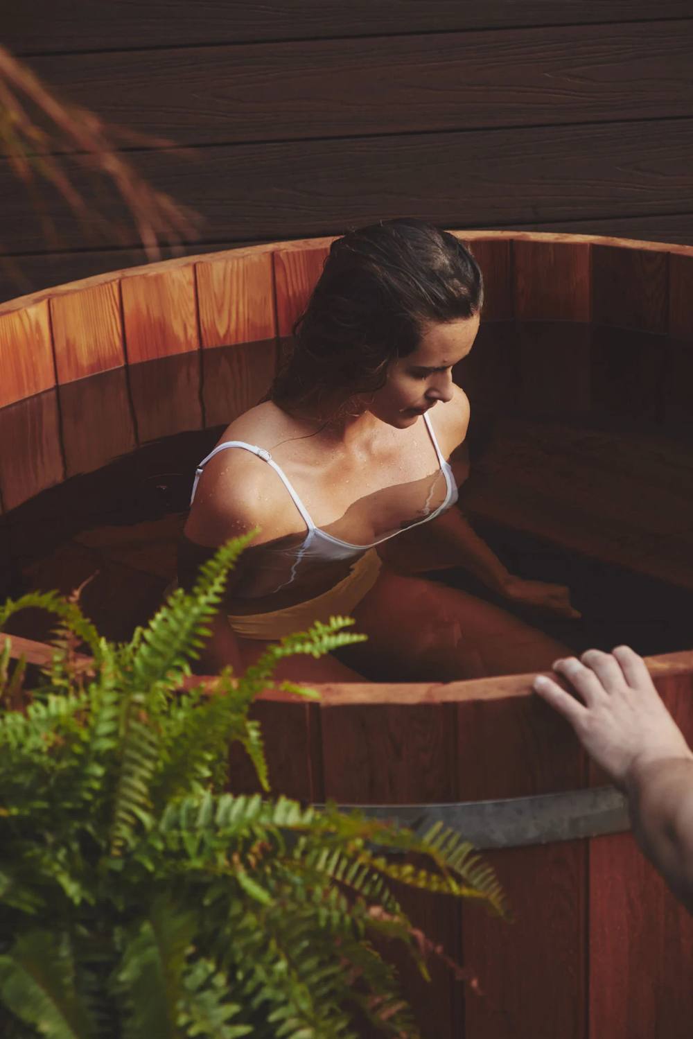 Woman soaking in a wooden cold plunge tub surrounded by ferns at ChoZen